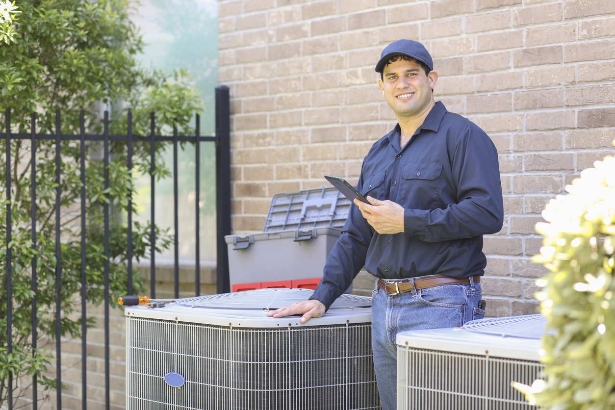 Smiling HVAC technician in uniform standing beside outdoor air conditioning units, holding a digital tablet during a maintenance inspection.